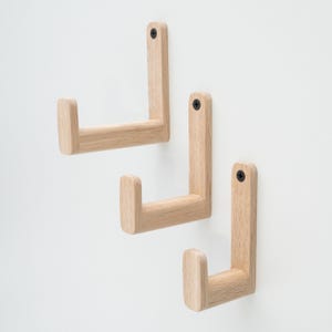 Może przedstawiać: Trzy drewniane haczyki ścienne w jasnym odcieniu drewna. Haczyki mają kształt litery L i są zamontowane na białej ścianie.