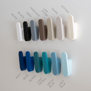 May include: A selection of wooden wall hooks in a range of colours: white, black, grey, hazelnut, lime stone, taupe, and cotton. Below are hooks in navy, blue, petrol, light petrol, arctic blue, and light blue. Colour names are printed on the wall.