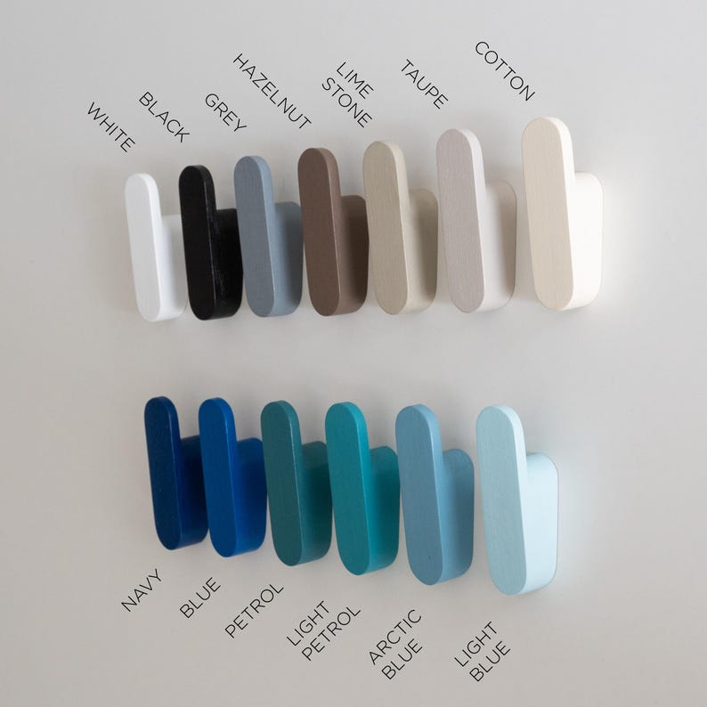 May include: A selection of wooden wall hooks in a range of colours: white, black, grey, hazelnut, lime stone, taupe, and cotton. Below are hooks in navy, blue, petrol, light petrol, arctic blue, and light blue. Colour names are printed on the wall.