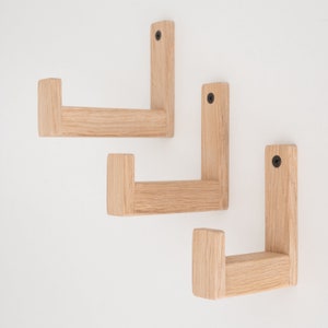 May include: Three wooden wall hooks with a natural finish are mounted on a white wall. Each hook has a rectangular backplate and a horizontal arm, with a black screw visible on each backplate. The hooks are evenly spaced.