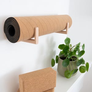 Op de afbeelding: Een opgerolde yogamat in lichtbruin hangt aan een houten houder aan een witte muur. Daaronder staat een kleine groene plant in een grijze pot op een witte plank, met twee bruine yogablokken.