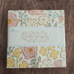 May include: A square stack of fabric squares with pinked edges, featuring a floral pattern in soft pink, yellow, and light blue on a white background. A paper band wraps around the center with the text Riley Blake Designs, little posies, Carla Naylor.