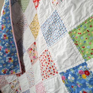 May include: A patchwork quilt featuring a grid of alternating white squares and various colorful patterned fabric squares. The patterns include floral prints in red, blue, yellow, and green, along with blue gingham and pink polka dots, finished with a striped border.