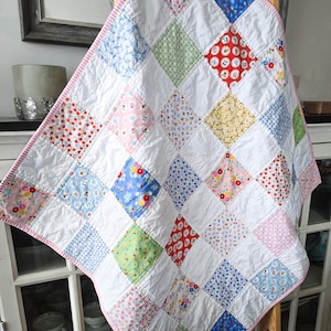 May include: A patchwork quilt featuring a repeating pattern of white squares alternating with squares of various floral and geometric prints in red, blue, yellow, pink, and green. The quilt is finished with a pink and white striped fabric binding.