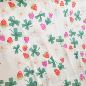 May include: White fabric with a repeating pattern of green four-leaf clovers, pink and red strawberries, and white daisies.