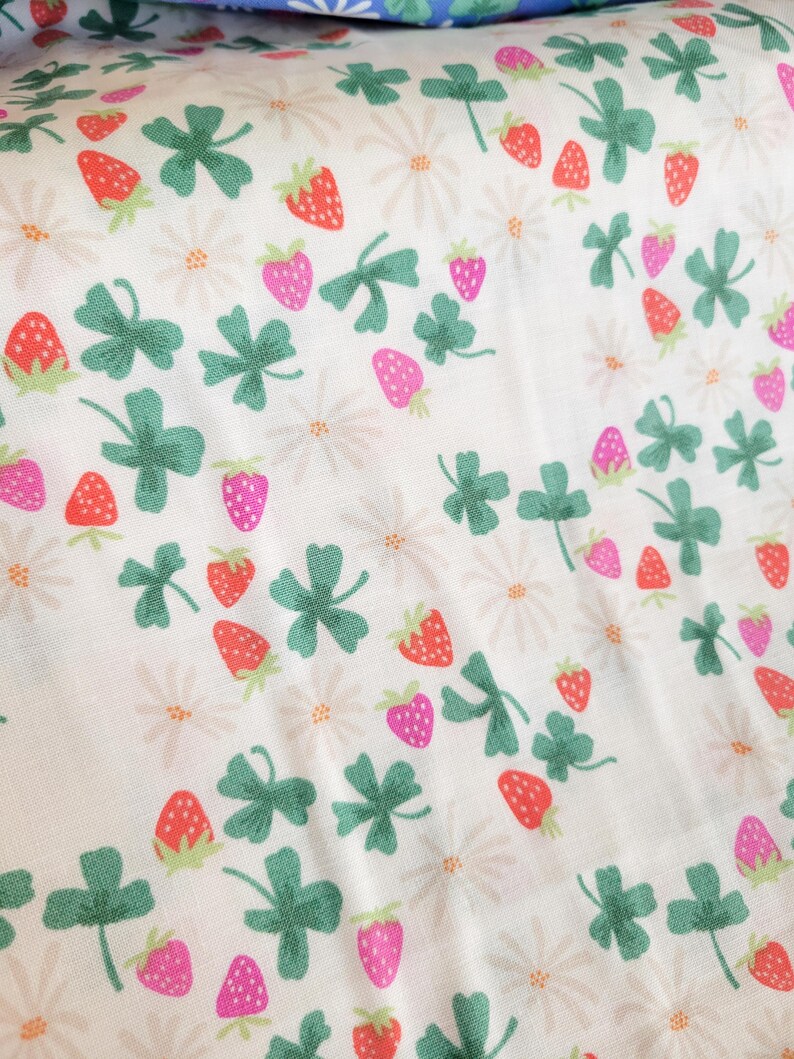 May include: White fabric with a repeating pattern of green four-leaf clovers, pink and red strawberries, and white daisies.