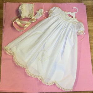 May include: White christening gown with lace trim and a matching bonnet. The gown has a scalloped lace trim and a small embroidered design on the front.