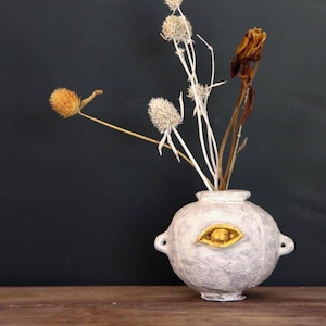 May include: A round, off-white vase with a gold eye-shaped accent, holding dried flowers. The vase has two small handles and sits on a wooden surface. The background is a dark, solid color.