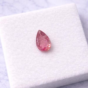 May include: A pear-shaped, pink gemstone rests on a white, textured surface. The gemstone has a translucent quality, with variations in color from light to deep pink. The background is a soft, marbled pattern.