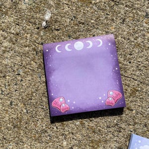 A purple sticky note with a moon and star motif is lying on a concrete surface.