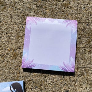 a square notepad with a purple border and a design of purple crystals on it, placed on a concrete surface.