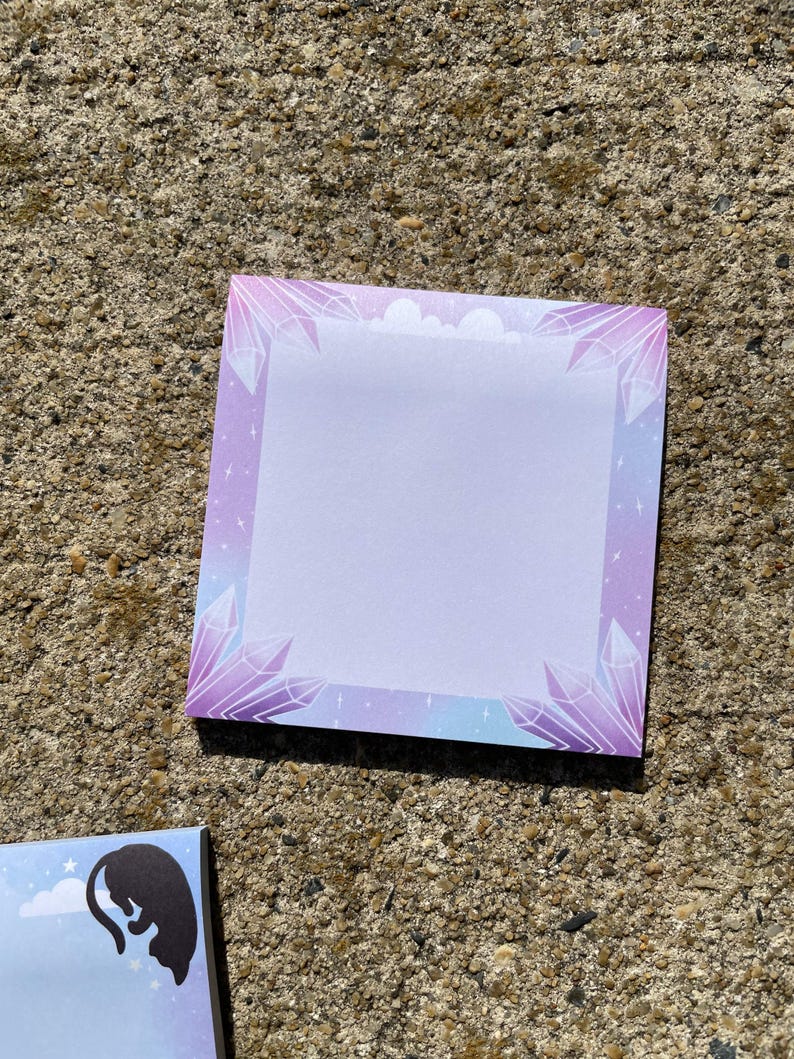 a square notepad with a purple border and a design of purple crystals on it, placed on a concrete surface.