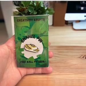 Könnte beinhalten: Eine grüne und weiße Karte mit dem Text "Creativity Erupts" und "Enamel Pin" in einem Kreis. Die Karte zeigt eine gelbe und schwarze Emaille-Anstecknadel mit einem Pied Ball Python. Der Text "Pied Ball Python" ist unter der Anstecknadel gedruckt.