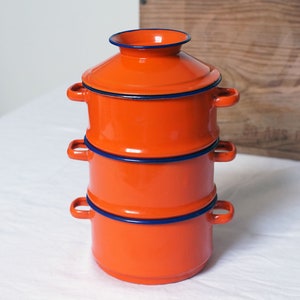 Peut inclure: Un ensemble de trois bols empilables en émail orange avec une bordure bleue. Les bols sont empilés les uns sur les autres, le plus grand en bas et le plus petit en haut.