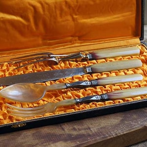 Puede incluir: Un juego de cinco cubiertos antiguos de plata plateada con mangos de nácar, presentados en una caja de madera forrada de terciopelo.