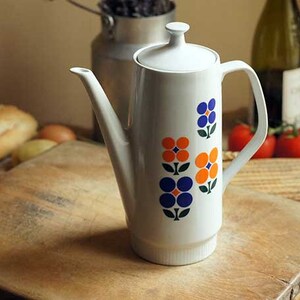 Peut inclure: Cafetière en céramique blanche avec un motif floral bleu, orange et vert. La cafetière a un couvercle et un bec verseur.
