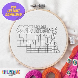 Pode incluir: Um aro de bordado branco com um contorno preto do estado de Nebraska e o texto "Let me just sneak on past ya" e "OPE" em negrito. O aro é cercado por fio de bordado colorido.