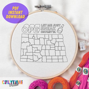 Könnte beinhalten: Ein weißer Stickrahmen mit einer schwarzen Umrandung des Bundesstaates North Dakota. Der Text "Let me just sneak on past ya" steht über der Umrandung des Bundesstaates. Neben dem Text ist eine kleine Flasche Alkohol gezeichnet. Der Rahmen ist von buntem Stickgarn umgeben.