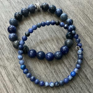 K&ouml;nnte beinhalten: Zwei geschichtete Armb&auml;nder mit runden Perlen in verschiedenen Blaut&ouml;nen, darunter Marineblau, K&ouml;nigsblau und Schiefergrau. Das Armband mit den gr&ouml;&szlig;eren Perlen enth&auml;lt eine einzelne silberfarbene, texturierte Metallperle als Zwischenelement.