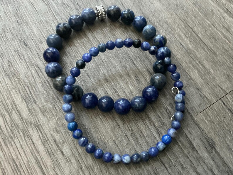 K&ouml;nnte beinhalten: Zwei geschichtete Armb&auml;nder mit runden Perlen in verschiedenen Blaut&ouml;nen, darunter Marineblau, K&ouml;nigsblau und Schiefergrau. Das Armband mit den gr&ouml;&szlig;eren Perlen enth&auml;lt eine einzelne silberfarbene, texturierte Metallperle als Zwischenelement.