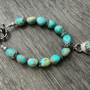 May include: A bracelet featuring polished turquoise beads with brown matrix patterns, interspersed with small silver-toned metal spacers and decorative beads. The piece includes a toggle clasp and a single oval pendant with a bezel-set turquoise stone.