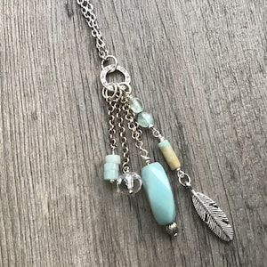 May include: Silver-toned necklace with a dangling design. The necklace features a series of small chains, beads in shades of blue, clear, and tan, and a silver feather charm. The necklace is displayed on a wooden surface.