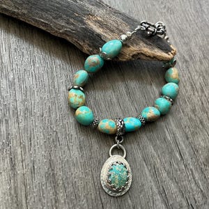 May include: A bracelet featuring smooth, rounded turquoise beads with brown matrix patterns, accented by small silver-toned metal spacers. A central oval pendant displays a matching turquoise stone set in a textured silver-toned metal frame.