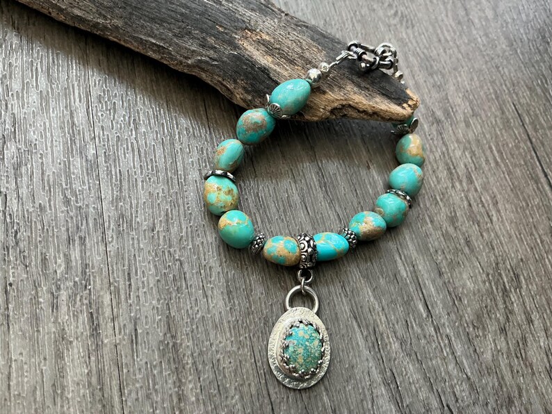 May include: A bracelet featuring smooth, rounded turquoise beads with brown matrix patterns, accented by small silver-toned metal spacers. A central oval pendant displays a matching turquoise stone set in a textured silver-toned metal frame.