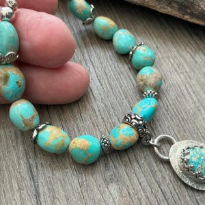 May include: A bracelet featuring polished, oval-shaped turquoise beads with brown matrix veining, interspersed with ornate silver-toned metal spacers and bead caps, finished with a circular silver-toned pendant containing a matching turquoise stone.