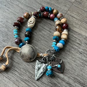 May include: A beaded bracelet with turquoise, brown, and cream-colored beads. The bracelet features a silver coin, an arrowhead charm, and a heart-shaped charm with a bull skull design. Tan leather ties are attached.