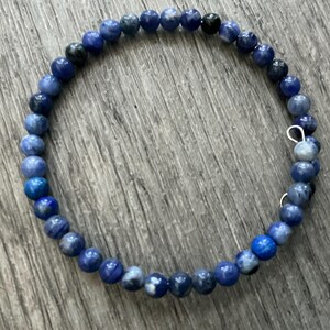 K&ouml;nnte beinhalten: Ein kreisf&ouml;rmiges Wickelarmband bestehend aus kleinen, runden, polierten Perlen in verschiedenen Blaut&ouml;nen, die von hellem Himmelblau bis zu tiefem Marineblau reichen, wobei einige Perlen gesprenkelte wei&szlig;e oder dunkelgraue Muster und eine kleine silberfarbene Metallschlaufe aufweisen.