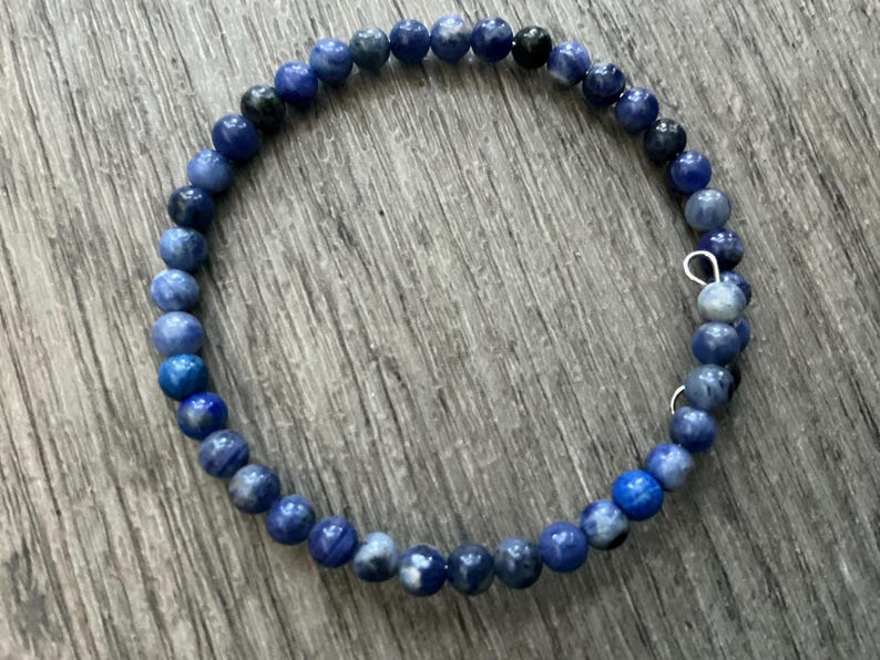 K&ouml;nnte beinhalten: Ein kreisf&ouml;rmiges Wickelarmband bestehend aus kleinen, runden, polierten Perlen in verschiedenen Blaut&ouml;nen, die von hellem Himmelblau bis zu tiefem Marineblau reichen, wobei einige Perlen gesprenkelte wei&szlig;e oder dunkelgraue Muster und eine kleine silberfarbene Metallschlaufe aufweisen.