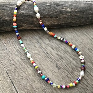 May include: A colorful beaded necklace with a variety of shapes and sizes. The necklace features white, oval-shaped beads interspersed with smaller, round, and irregularly shaped beads in shades of red, blue, green, and yellow. The necklace is displayed on a wooden surface.