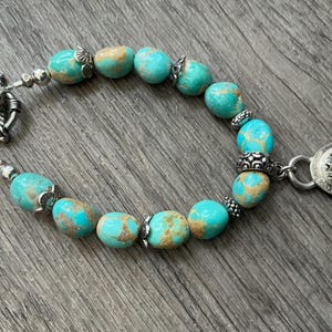 May include: A bracelet featuring smooth, rounded turquoise beads with brown matrix patterns, interspersed with small silver metal spacers and decorative caps. The piece includes a silver toggle clasp and a single oval turquoise pendant charm.