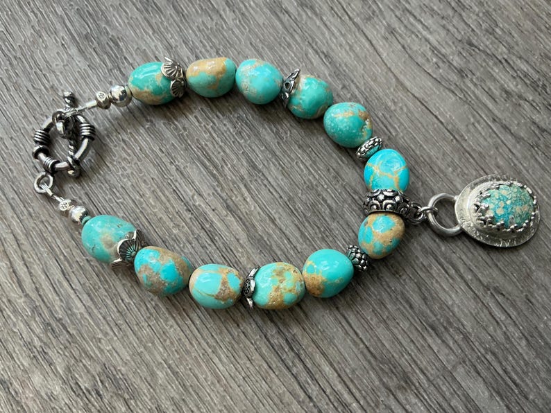 May include: A bracelet featuring smooth, rounded turquoise beads with brown matrix patterns, interspersed with small silver metal spacers and decorative caps. The piece includes a silver toggle clasp and a single oval turquoise pendant charm.