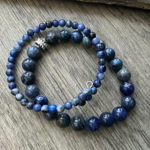 K&ouml;nnte beinhalten: Ein Wickelarmband mit zwei verschiedenen Str&auml;ngen aus blauen und dunkelgrauen runden Perlen. Ein Strang besteht aus kleineren, gleichm&auml;&szlig;igen Perlen, w&auml;hrend der andere gr&ouml;&szlig;ere, variierende Perlen und eine silberfarbene, strukturierte Metallperle enth&auml;lt.