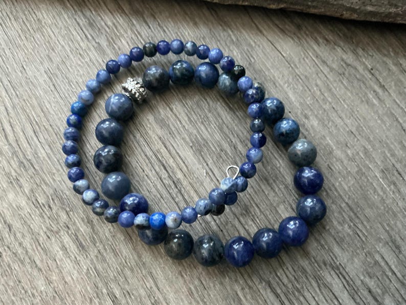 K&ouml;nnte beinhalten: Ein Wickelarmband mit zwei verschiedenen Str&auml;ngen aus blauen und dunkelgrauen runden Perlen. Ein Strang besteht aus kleineren, gleichm&auml;&szlig;igen Perlen, w&auml;hrend der andere gr&ouml;&szlig;ere, variierende Perlen und eine silberfarbene, strukturierte Metallperle enth&auml;lt.