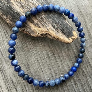 K&ouml;nnte beinhalten: Ein kreisf&ouml;rmiges Armband aus kleinen, polierten, kugelf&ouml;rmigen blauen Perlen in verschiedenen Nuancen von Marineblau, K&ouml;nigsblau und Hellblau, mit einem kleinen Verschluss aus silberfarbenem Metalldraht am unteren Ende.