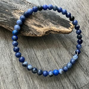 K&ouml;nnte beinhalten: Ein kreisf&ouml;rmiges Armband aus kleinen, runden, polierten Perlen in verschiedenen Blaut&ouml;nen, die von hellem Himmelblau bis zu tiefem Marineblau reichen, wobei einige Perlen dunkelgraue oder schwarze Einschl&uuml;sse und zwei kleine silberfarbene Metallringe aufweisen.