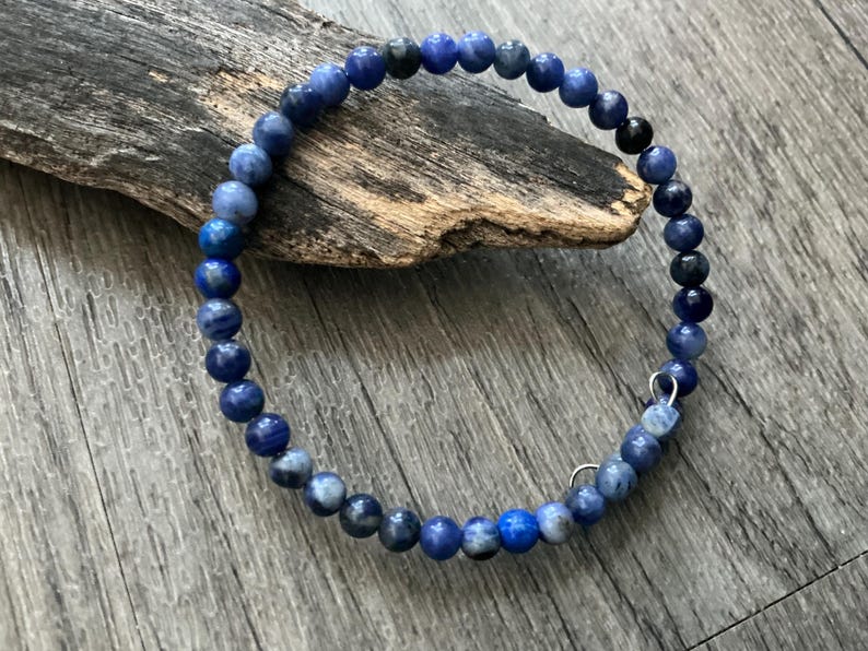K&ouml;nnte beinhalten: Ein kreisf&ouml;rmiges Armband aus kleinen, runden, polierten Perlen in verschiedenen Blaut&ouml;nen, die von hellem Himmelblau bis zu tiefem Marineblau reichen, wobei einige Perlen dunkelgraue oder schwarze Einschl&uuml;sse und zwei kleine silberfarbene Metallringe aufweisen.