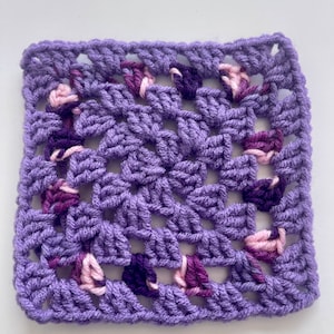 Puede incluir: Un cuadrado de ganchillo morado con un patrón geométrico. El cuadrado está hecho con una variedad de tonos de hilo morado, incluyendo rosa y morado oscuro.