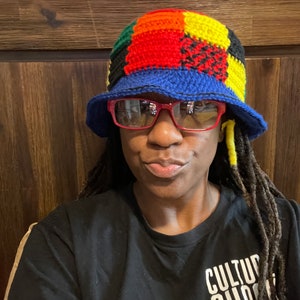 May include: A person wearing a colorful crocheted bucket hat with a blue brim and red sunglasses. The hat has a patchwork design with squares of red, yellow, green, and black yarn. The person is wearing a black t-shirt with the text "CULTURE SHOCK SAN DIEGO" printed on it.