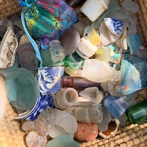 May include: A woven basket filled with sea glass, pottery shards, and small glass bottles. A teal drawstring bag with a colorful floral pattern sits among the contents. The sea glass pieces are various colors, including green, blue, and clear.