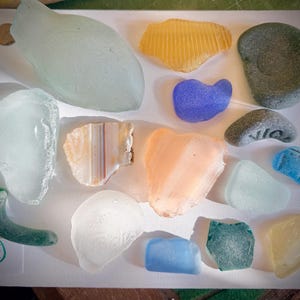 May include: A collection of sea glass pieces in various shapes and colors, including blue, green, yellow, and clear. Some pieces have a frosted appearance, while others are smooth. A small coin and a piece of paper with the number 3 are also visible.