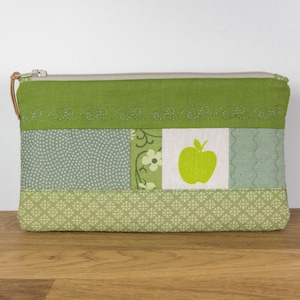 Könnte beinhalten: Ein grüner Patchwork-Reißverschlussbeutel mit Apfelmuster. Der rechteckige Beutel hat verschiedene Grüntöne mit unterschiedlichen Mustern, darunter Punkte und Blumendrucke. Auf einem weißen Quadrat ist ein hellgrüner Apfel aufgedruckt.