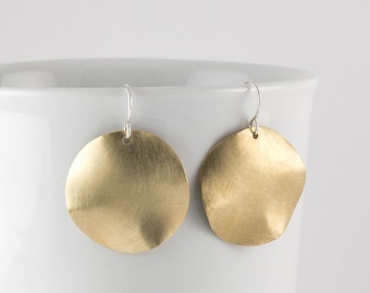 May include: A pair of gold-toned brass earrings with a hammered finish. The earrings are shaped like a circle with a slightly wavy edge. The earrings are hanging from silver hooks.