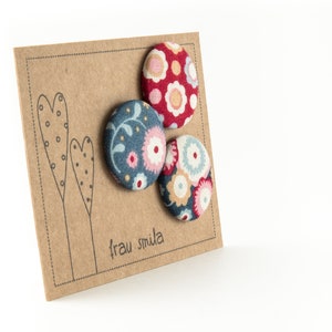 Puede incluir: Tres botones cubiertos de tela con motivos florales. Los botones est&aacute;n en una tarjeta marr&oacute;n con el texto "frau smula" impreso en ella.