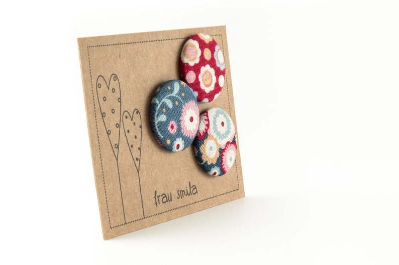 Puede incluir: Tres botones cubiertos de tela con motivos florales. Los botones est&aacute;n en una tarjeta marr&oacute;n con el texto "frau smula" impreso en ella.