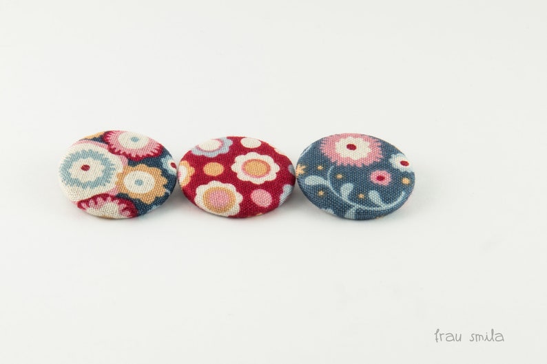 Puede incluir: Tres botones cubiertos de tela con motivos florales. Los botones son en tonos de azul, rojo y rosa.