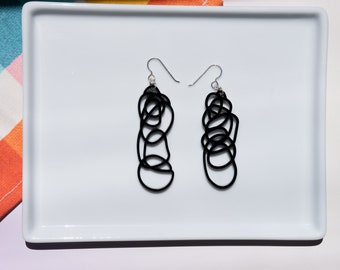 Puede incluir: Un par de pendientes negros colgantes con un diseño abstracto único. Los pendientes están hechos de alambre negro delgado, retorcido y anudado para crear un patrón geométrico.